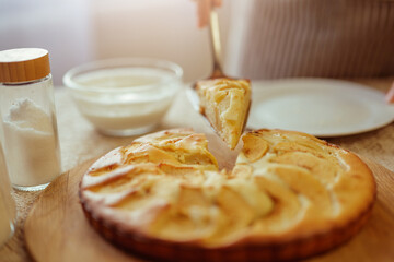 A cook in a striped apron lifts a slice of freshly baked apple pie with a spatula, with the rest of the pie and baking ingredients on the table.