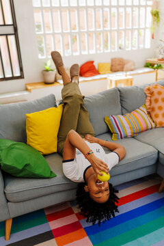 Woman lying upside down on sofa eating apple in modern apartment