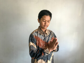 A teenage boy wearing a batik shirt doing a respectful greeting with hands together, isolated on white
