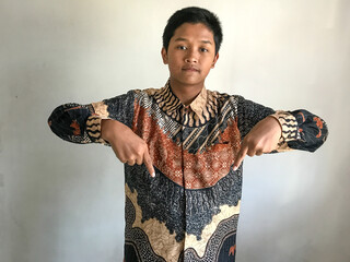 A teenage boy wearing a batik shirt is pointing both of his index fingers downward, isolated on white