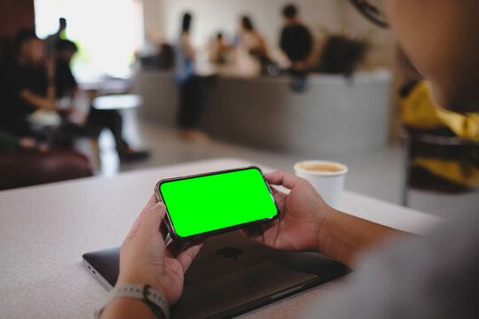 Person holding smartphone with green screen in landscape orientation at cafe table with coffee cup and laptop in background, ready for video or game display.