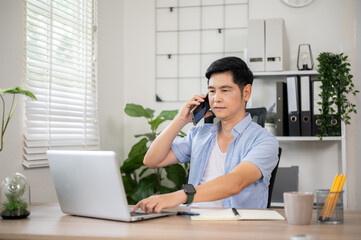 Asian man talking on phone and typing on laptop sitting at wooden table in office or working room.