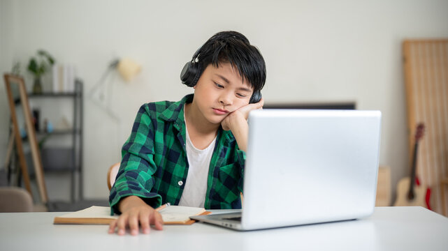 Asian kid elementary school boy wearing headphone looking at laptop on table with hand support head