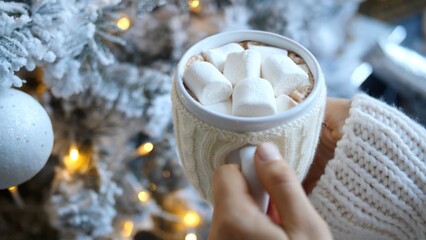 Woman wearing white woolen sweater is holding a mug of hot chocolate with marshmallows near decorated christmas tree with lights and white ornament