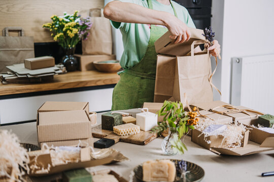 Gift boxes with handmade soaps are prepared on a kitchen table while a person ties a kraft bag. DIY business visuals, handmade product packaging, entrepreneur life, craft fair prep