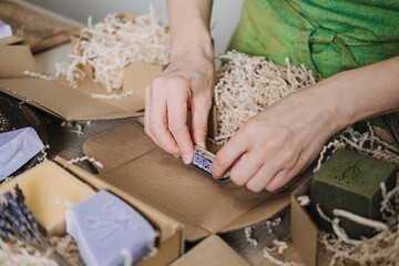 Person stamps kraft soap box during packaging surrounded by lavender bars and natural filler. Product customization, handcrafted appeal, soap maker branding, creative product identity