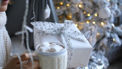 Woman wearing a white sweater is enjoying a mug of hot chocolate with marshmallows and holding a christmas gift wrapped in white paper with a silver ribbon near a decorated christmas tree with lights