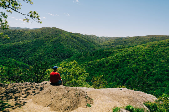 Man in red shirt sits on cliff overlooking Blue Ridge Mountains forest - Powered by Adobe