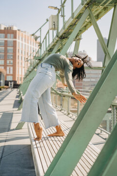 Woman arches back in dramatic pose on bench beneath city bridge