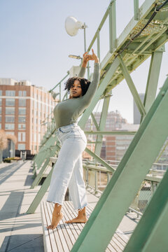Woman poses barefoot on bench, arms raised, under city bridge beams