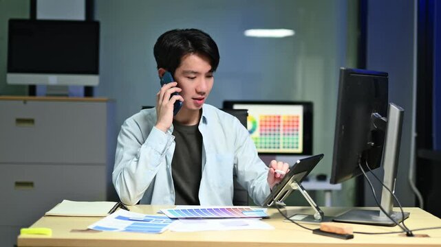 UX UI designer working late in a modern office, multitasking with phone and tablet. App wireframes on screen show tech, focus, and mobile innovation.
