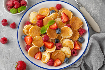 Tiny pancakes with fresh berries, honey and mint for breakfast in a bowl with a cup of coffee on a gray concrete background.