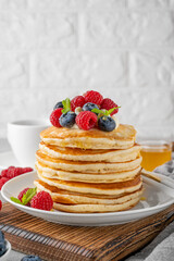 A stack of pancakes with whipped cream, fresh berries and honey on a white plate on a gray concrete background.