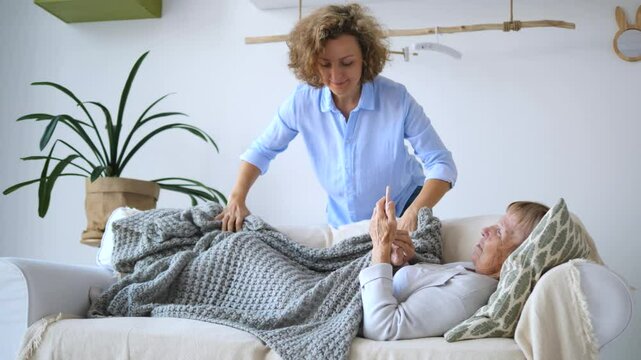 Caring daughter covering her elderly mother with a warm blanket while she rests on a comfortable sofa in the living room, demonstrating love and support for her aging parent