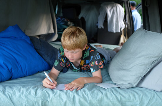 Boy writing postcards while lying on a bed inside a van