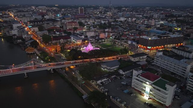 Night aerial of Surat Thani City Pillar Shrine glowing, with city lights, Tapi River, and traffic illuminating the urban landscape of Thailand.
