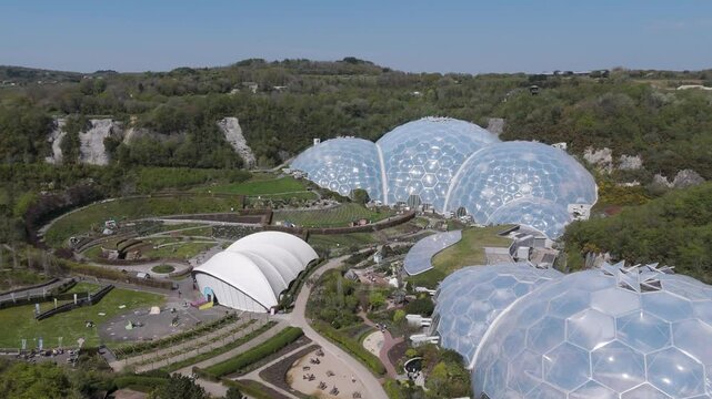 Broad aerial panorama of Eden Project dome clusters shimmering above concentric gardens and forested slopes, illustrating regeneration, biodiversity and renewable innovation