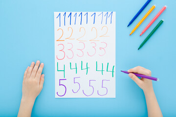 Little child boy hand holding color pen and writing colorful different numbers on white paper on blue table background. Pastel color. Doing school exercise. Closeup. Point of view shot. Top down view.