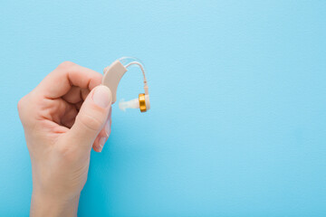 Young adult woman hand fingers holding and showing hearing aid on light blue table background. Pastel color. Closeup. Empty place for text. Top down view.