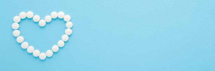 Heart shape created from white pills on light blue table background. Pastel color. Closeup. Healthcare concept. Wide banner. Empty place for text. Top down view.
