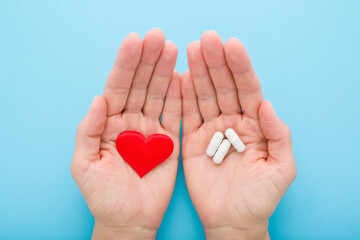 Young adult woman opened palms holding white capsule pills and red heart shape on light blue table background. Closeup. Pastel color. Receiving medicaments. Point of view shot. Top down view.