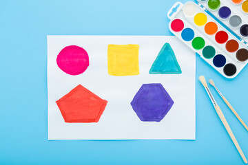 Colorful watercolor palette, paintbrushes and drawn colorful geometric shapes on white paper on blue table background. Pastel color. Creativity different forms learning for little kids. Top down view.