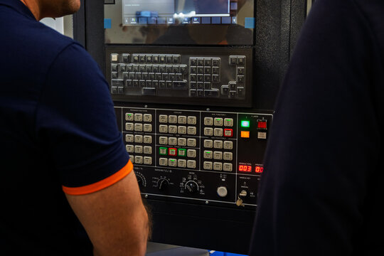 Two people stand together next to an industrial machine using a panel with many buttons