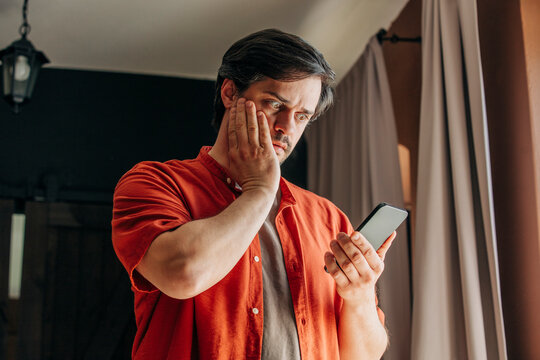 Man in red shirt looking at smartphone with horror, reacting to financial crisis and falling stocks indoors