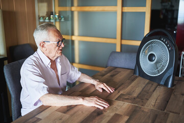 Senior man cooling down with electric fan at home during heat wave