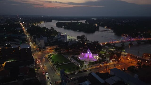 Night aerial of Surat Thani City Pillar Shrine glowing, with city lights, Tapi River, and traffic illuminating the urban landscape of Thailand.