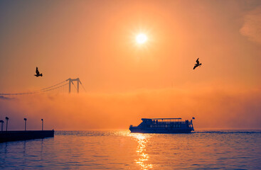 Fototapeta premium Ortakoy Mosque and Bosphorus Bridge in Istanbul, Turkey. Dramatic sky. The 15 July Martyrs Bridge in a fog. Passenger Boat on the Sea on a Foggy Day.