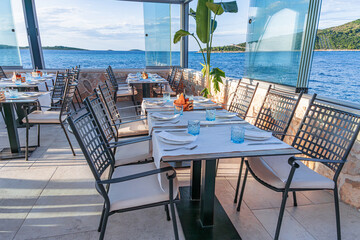 Tranquil seaside restaurant with modern wooden tables and chairs arranged under large umbrellas along a scenic waterfront promenade.