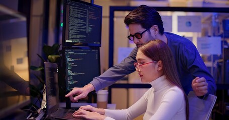 Two Asian software engineers collaborate at night in modern office, writing machine learning algorithms and debugging artificial intelligence code on multiple monitors in AI tech development workflow.