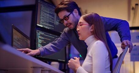 Two Asian software engineers collaborate at night in modern office, writing machine learning algorithms and debugging artificial intelligence code on multiple monitors in AI tech development workflow.