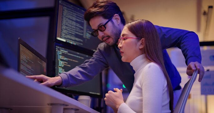 Two Asian software engineers collaborate at night in modern office, writing machine learning algorithms and debugging artificial intelligence code on multiple monitors in AI tech development workflow.