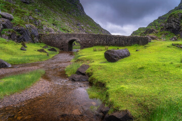 A picturesque view showcasing a historic stone Wishing Bridge elegantly spanning across a gentle stream, beautifully surrounded by vibrant greenery of Gap of Dunloe and dramatic skies, Kerry, Ireland