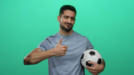 Young man holding soccer ball showing thumbs up against isolated green wall background in a confident and charming pose suggesting positivity and sportsmanship.