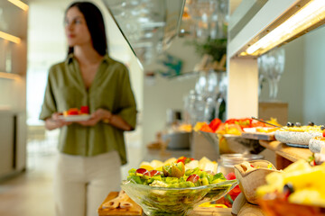 Fresh fruit salad in focus on a hotel breakfast buffet, with a blurred woman holding a plate in the background