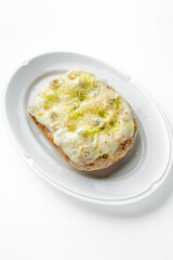 A slice of bread with melted cheese, garnished with green herbs and seasoning, served on a white oval plate. The dish looks appetizing and well-styled, perfect for food photography