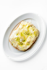 A slice of bread with melted cheese, garnished with green herbs and seasoning, served on a white oval plate. The dish looks appetizing and well-styled, perfect for food photography