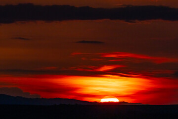 Fiery sun setting below the horizon with glowing red clouds above the hills. A powerful visual of nature’s final light of day.