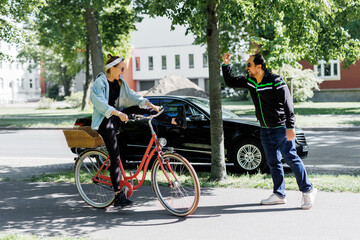 Man gestures angrily toward woman riding red bicycle on sidewalk, suggesting conflict in shared urban space. Scene shows tension between cyclist and pedestrian under trees on sunny day