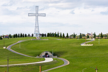 A monumental steel cross stands tall over green hills, with religious mosaic at its base and cloudy sky above — symbol of faith and national pride