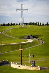 Panoramic view of a religious hilltop site with monumental cross and underground chapel entrance. Pilgrims walk toward the sacred location