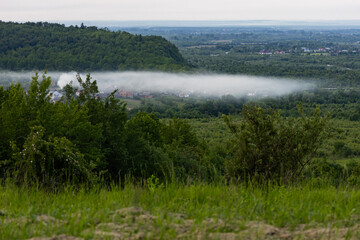 Low morning fog blankets a quiet village surrounded by green hills and forests. Peaceful countryside landscape captured from above