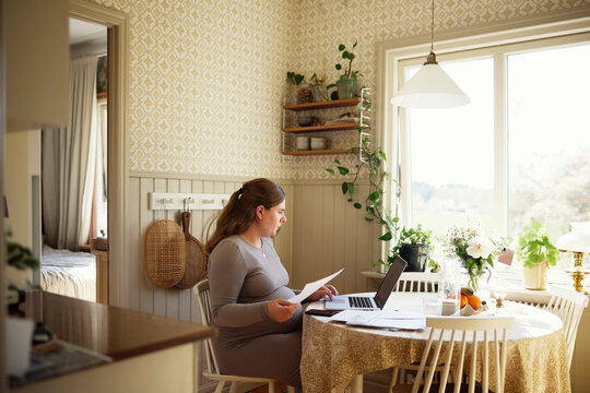 Pregnant woman using laptop while calculating bills sitting at table in home