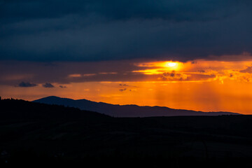 A mesmerizing gradient of fiery light and looming darkness unfolds as the sun hides behind rain-laced clouds over the Carpathian hills — a vivid capture of the storm-kissed twilight hour