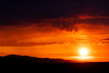 Bright golden sun hangs low over shadowed hills as vivid orange light paints the sky in waves of color and warmth