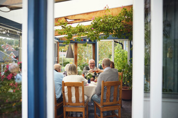 Male and female friends eating lunch and clinking beer glasses while sitting at dining table seen through patio door