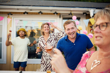 Happy man holding glass of alcohol drink dancing with friends in back yard during social gathering event